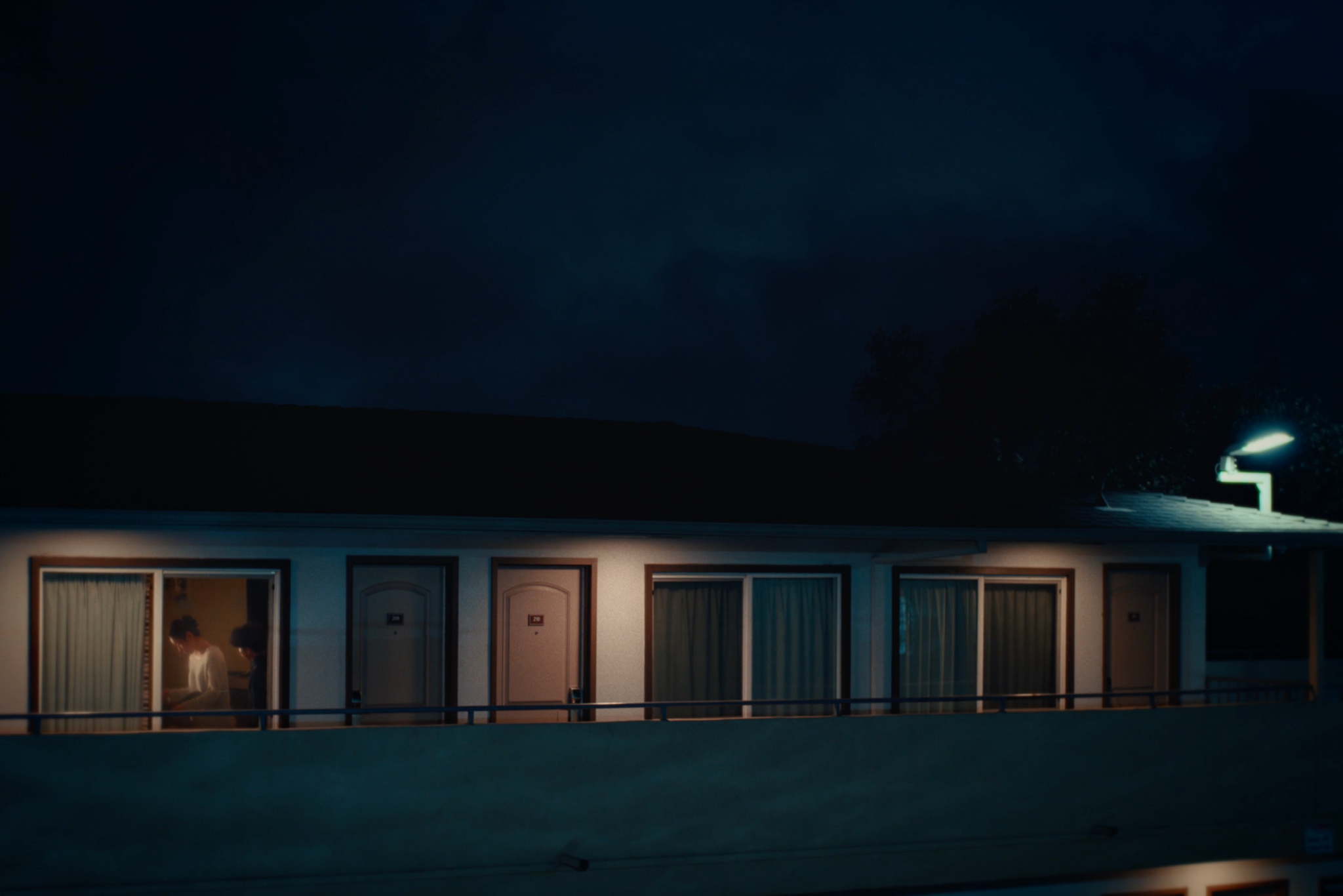 Night exterior of an apartment walkway from Sweet Boy, with two figures visible through a window